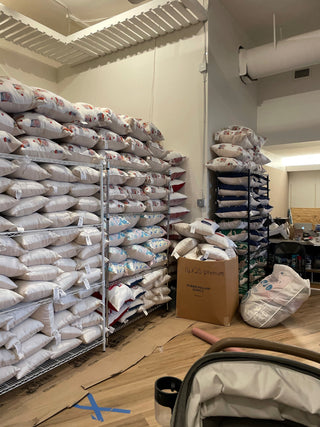 Shelves stacked with large bags of rice are lined against the wall in a room. Cardboard boxes, open bags, and various packing supplies are scattered on the wooden floor. The setting appears industrial or warehouse-like.