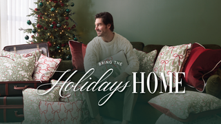 A man in a white sweater sits on a couch with festive pillows, next to a Christmas tree and gifts. Text reads: Bring the Holidays Home.