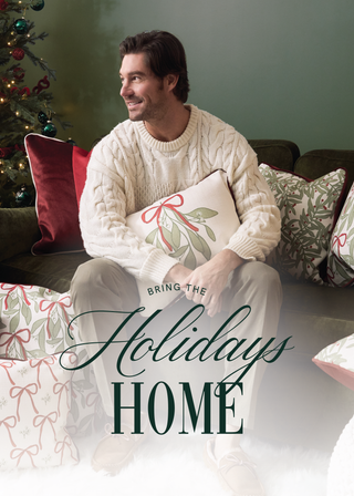 A man in a cream sweater and slippers sits on a sofa with festive pillows decorated with red bows and greenery, near a Christmas tree. Text reads, “Bring the Holidays Home.”.
