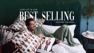 A smiling man in sage green pajamas lounges on a bed surrounded by holiday pillows with nutcracker designs. Text reads, Cozy up to our best selling holiday pillows. A mug with a matching nutcracker design sits nearby.