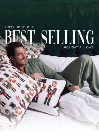 A smiling man in green pajamas lounges on a white bed, surrounded by decorative holiday pillows featuring nutcracker designs. Text reads: Cozy up to our best selling holiday pillows.