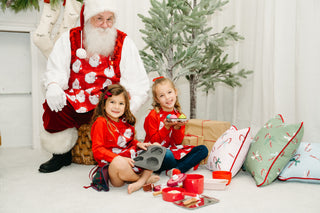 Two young girls in red outfits smile by a small Christmas tree with gifts, play food, and festive pillows. Behind them, someone poses as Santa Claus wearing the Childrens Santa Apron for the photo.