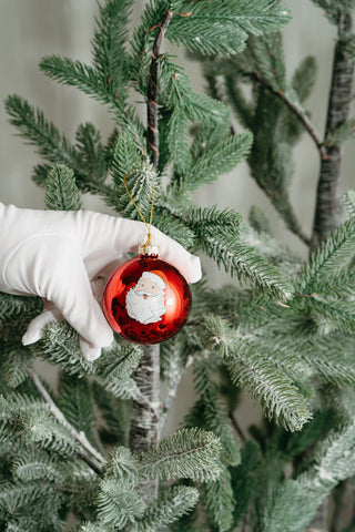 A gloved hand holds a Santa Globe Ornament (available in single, 2 or 4 pack) with a festive Santa illustration, ready to hang on your green artificial tree for a cheerful coastal holiday touch.