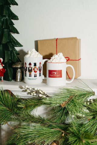 Two Nutcracker Coffee Mugs filled with hot chocolate and whipped cream sit on a mantel adorned with pine garland, with a wrapped holiday gift and a decorative tree in the background.