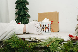 A gloved hand holds the Nutcracker Coffee Mug filled with whipped cream, set beside a wrapped holiday gift, pine garland, and a festive Christmas tree on a white mantel.