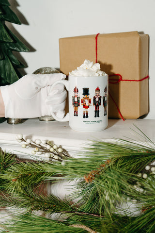 A gloved hand holds the Nutcracker Coffee Mug filled with hot chocolate and whipped cream, surrounded by a wrapped holiday gift, artificial pine branches, and festive décor for a cozy seasonal vibe.