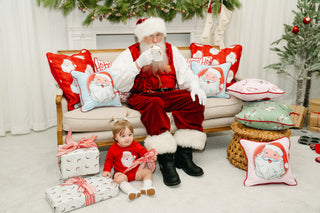Santa Claus relaxes on a festive couch with a mug, surrounded by the Santa Holiday Pillow Collection and the Childrens Santa Pillow in Baby Blue. Nearby, a child in red sits amid wrapped gifts, enhancing the joyful holiday decor.