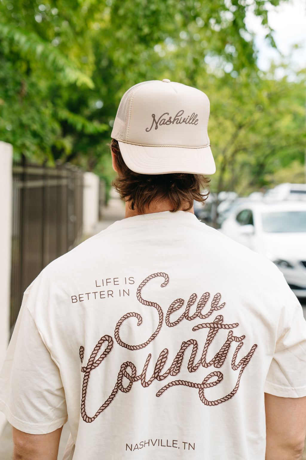 A man adjusts his SDS Nashville Rope Trucker Hat, complete with an adjustable snapback. He sports a cream T-shirt showcasing a cowboy hat graphic on the pocket. The softly blurred trees and building wall in the background accentuate his relaxed style.