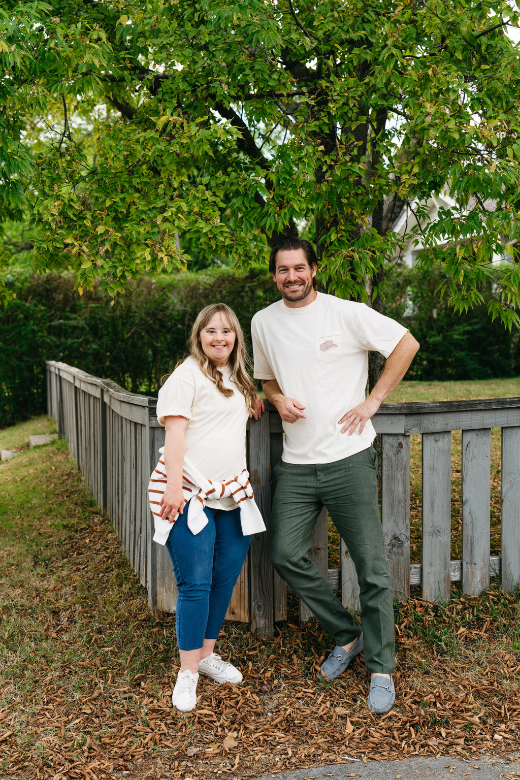 A person with light brown hair wears a beige Nashville cap and a white unisex Life is Better in Sew Country T-shirt. Greenery and a white wooden structure create an Americana backdrop.