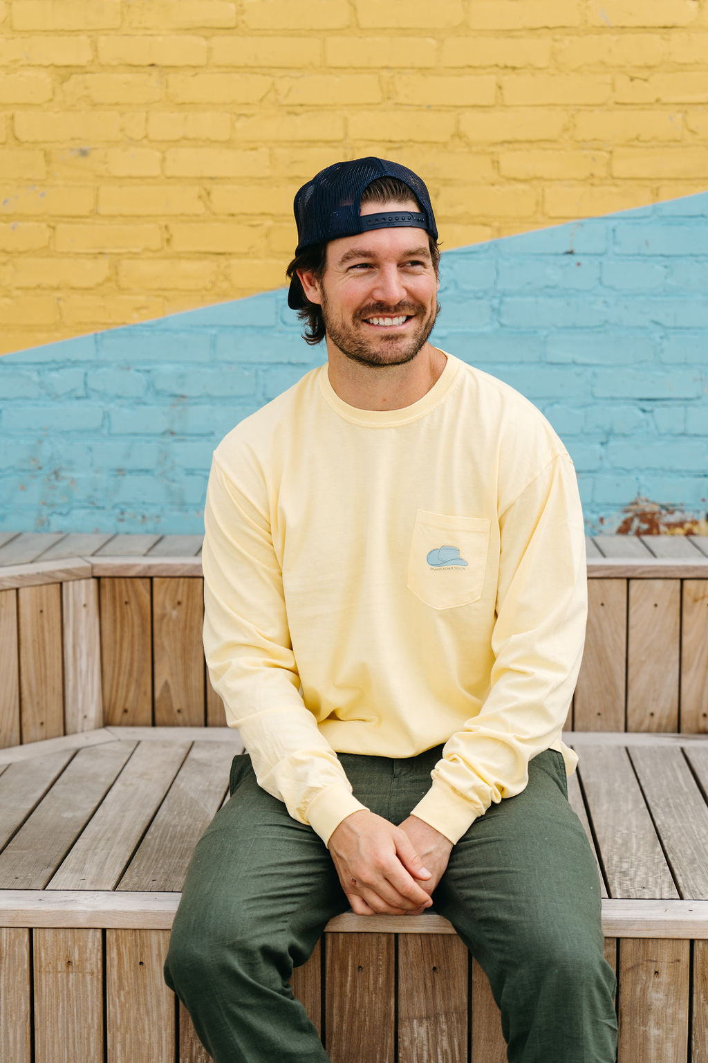 A man in a backward baseball cap and a yellow Life is Better in Sew Country Long Sleeve Tee relaxes on a wooden bench. A yellow and blue brick wall, echoing Nashvilles charm, provides the vibrant backdrop with Life is better in Sew Country and Nashville, TN printed on the shirt.