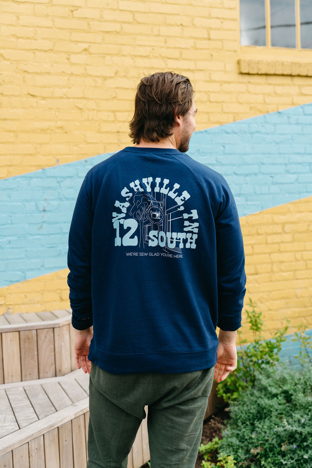 Wearing a SDS Nashville 12 South Crewneck Sweatshirt and a navy cap, a man sits thoughtfully on a wooden bench against the lively yellow and blue bricks of a Charleston storefront, adding charm to his contemplative pose.