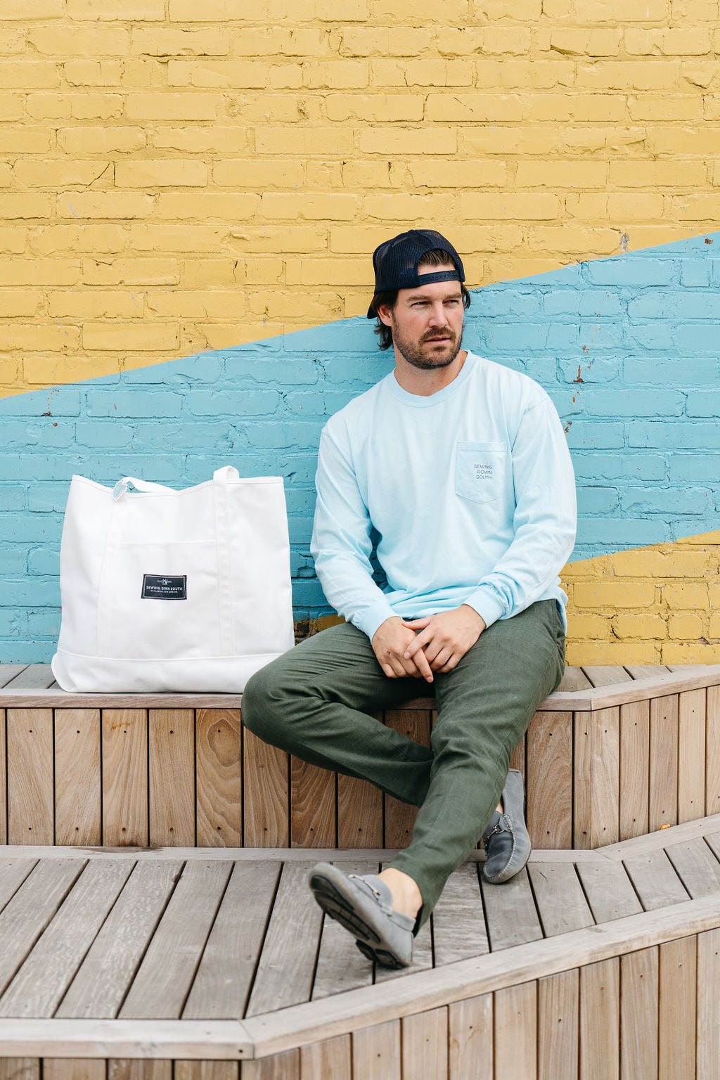 Facing away, a person wears the SDS Nashville Rope Long Sleeve Shirt, a unisex blue chambray tee made from 100% ring-spun cotton. It features Nashville script and Were sew glad youre here. They pair it with a cap displaying Nashville on the back, standing by a colorful wall.