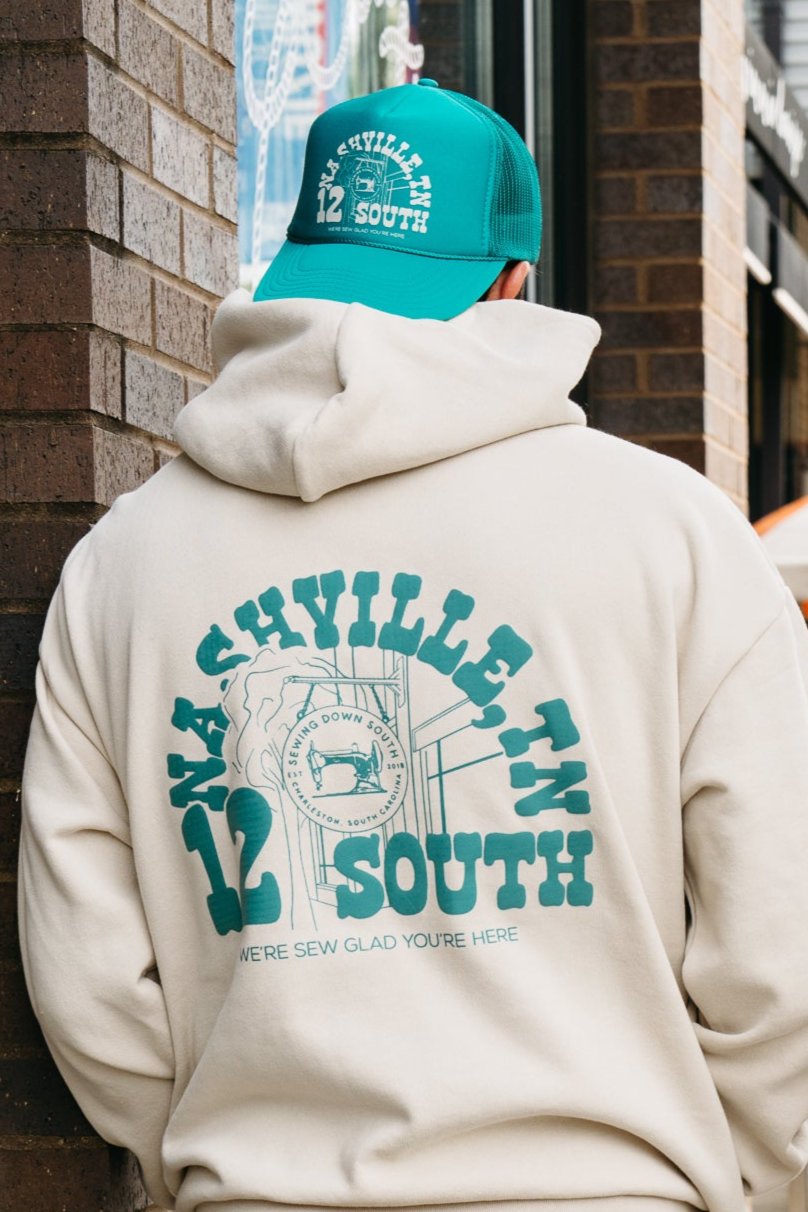 A person wearing an SDS Nashville 12 South Trucker Hat in turquoise and a white hoodie strolls down the street, framed by blurred signs and buildings.