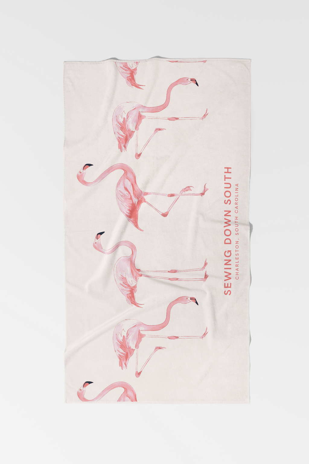 A sunhat, a magazine titled “REALTY,” and the Flamingo Beach Towel with pink flamingo prints are laid out on the sandy beach. The cotton towel has some sand on it.