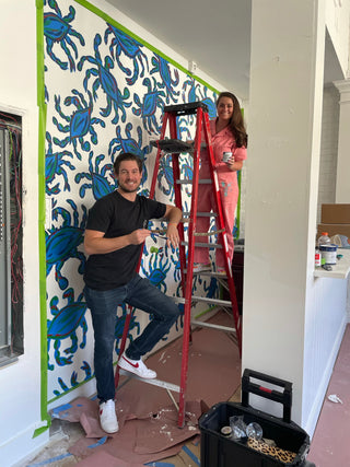 A man stands next to a red ladder while a woman in pink overalls stands on it, both smiling in front of a colorful, blue-patterned accent wall. Painting supplies and tools are scattered around them.