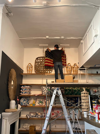 A person stands on a shelf hanging an American flag on a wall in a shop, surrounded by decorative items, shelves with textiles, a ladder, and various home goods.