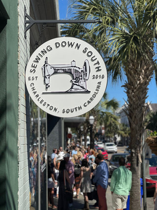 A round sign with a sewing machine illustration reads Sewing Down South, Est. 2018, Charleston, South Carolina. A crowd gathers outside the shop on a sunny street lined with palm trees.