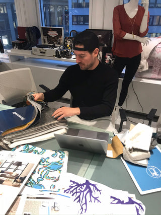 A man wearing a black shirt and cap sits at a desk, looking through fabric samples. The desk is covered with swatches, papers, and a laptop. A mannequin and shelves with bags are visible in the background.