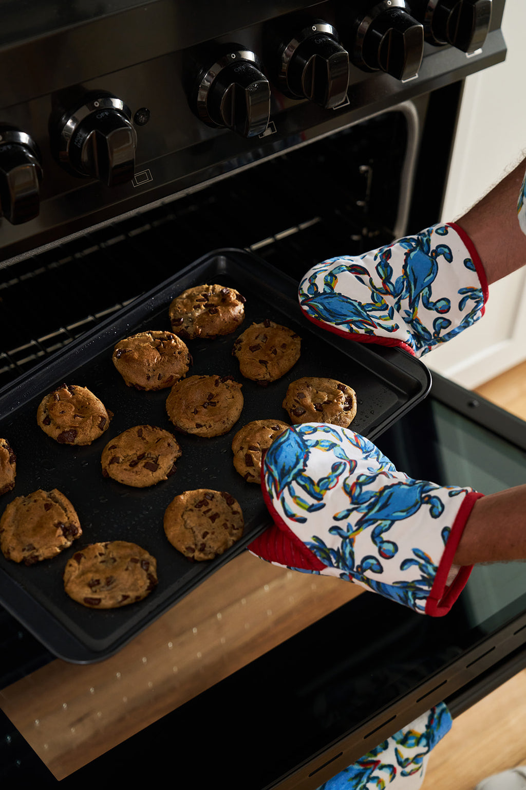 A person using playful Crab Craze Mini Oven Mitts with silicone grips takes a tray of freshly baked cookies from the oven, smiling in a charming kitchen adorned with a bouquet of white flowers.