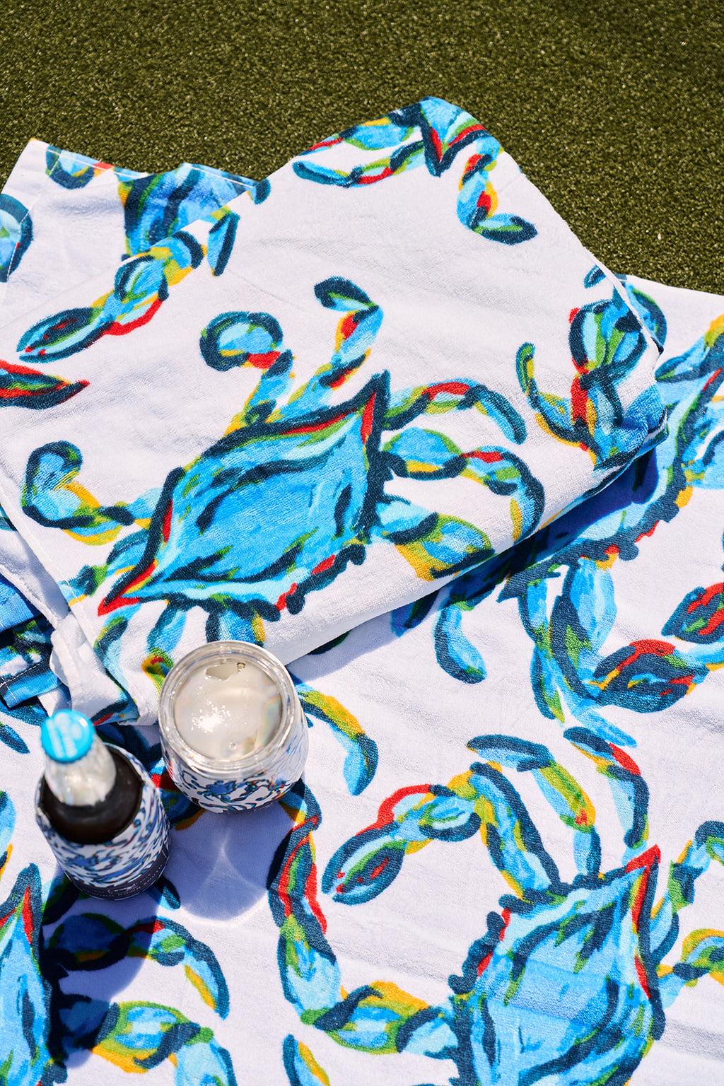 A man in sunglasses and a cap sits by the pool, smiling with a colorful Crab Craze Beach Towel from Sewing Down South draped around his shoulders. Hes sporting a blue shirt and shorts, with decorative pillows and an umbrella in the background.