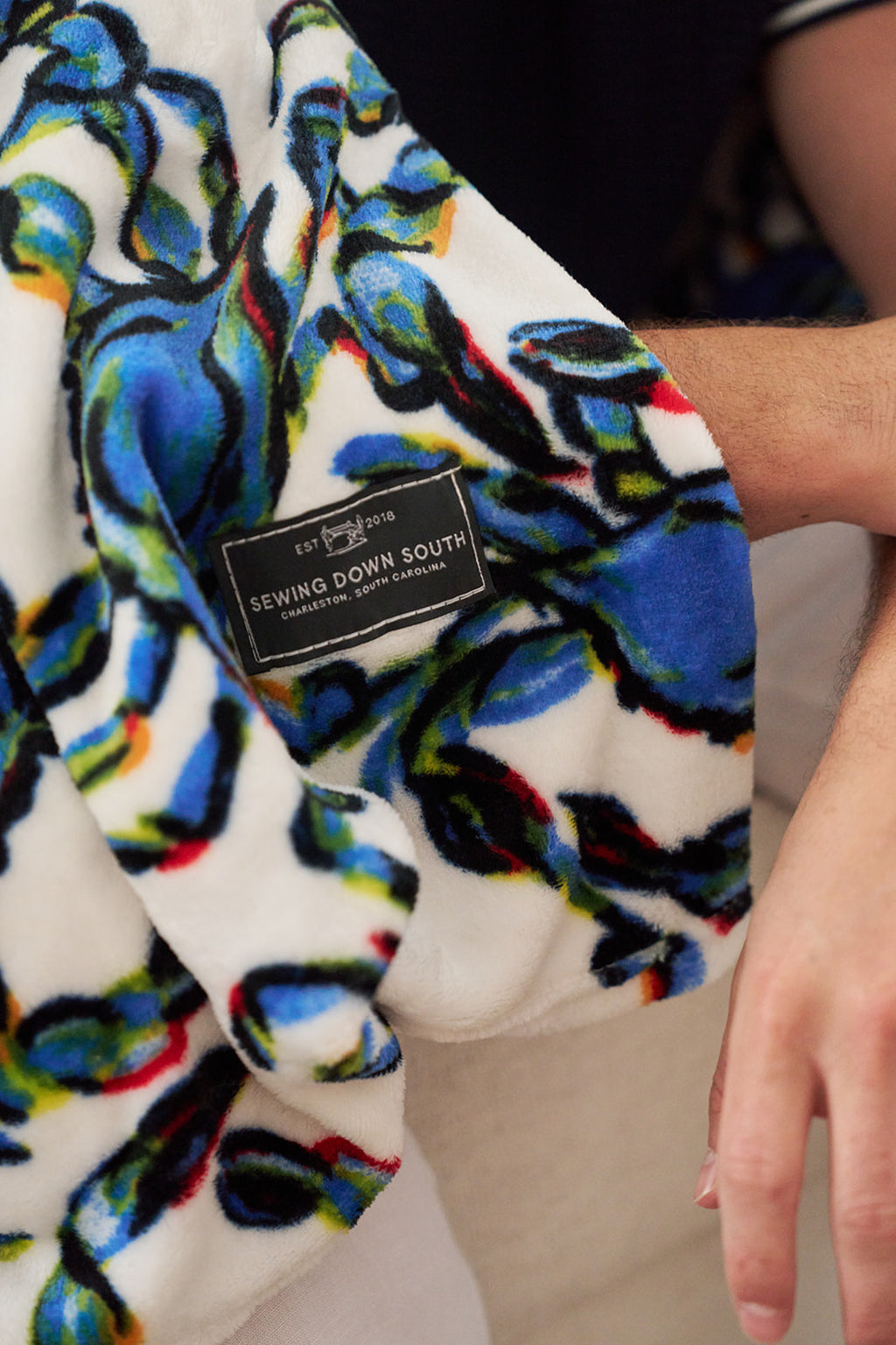 A man smiles while sitting on a couch, wrapped in a Crab Craze, Shades of Blue Blanket. Wearing a navy shirt with a striped collar and white pants, he embodies coastal chicness. The cozy setting enhances the relaxed vibe.