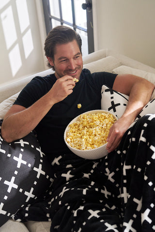 A man smiles and eats popcorn on a couch draped with The Paige Collection: Exes in Black Blanket, as sunlight streams through the window, creating a cozy setting.