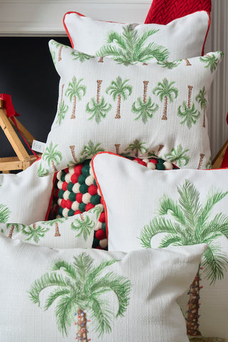A stack of white throw pillows, featuring the Holiday Palmetto Lumbar Pillow with green palm prints and red piping, is arranged in front of a colorful knit blanket for a cozy, festive look.