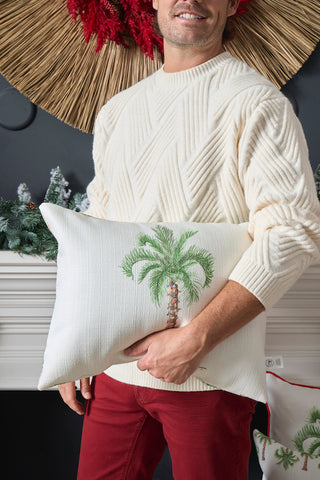 A man in a cream knit sweater and red pants holds the Holiday Palmetto Lumbar Pillow, featuring an embroidered palm tree. Festive holiday decorations with greenery and red accents enhance the cheerful setting.