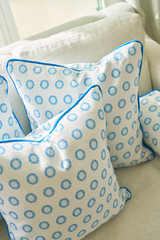 Three white throw pillows with blue sunburst designs and bright blue piping rest on a light sofa. One is the Calico Mini Floral Oversized Pillow, featuring a subtle mini floral pattern. Sunlight streams through the nearby window, brightening the scene.