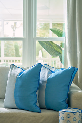 Two Spring Blue Blocks Oversized Pillows rest on a beige sofa by a large window with sheer curtains, as sunlight brightens a green plant and brings fresh, modern home décor vibes to the room.