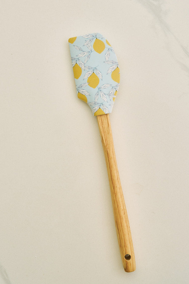 On a marble countertop, a recipe card sits beside a bowl of lemons. The heat-resistant Lots of Lemons Spatula, made from BPA-free silicone, rests on a lemon-themed towel. Nearby, lemon bars garnished with lemon slices and mint sit on a plate.