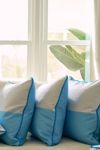 The Spring Blue Blocks Oversized Pillow and two other blue and white throw pillows are arranged on a light-colored couch by a sunlit window, adding a calming modern touch with green leafy plants in the background.