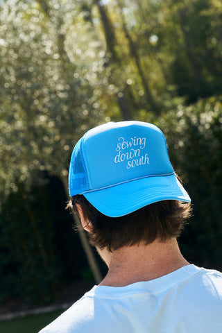A person with short brown hair wears The Siren Collection: Sewing Down South Trucker hat, embroidered with the brands name. They face away in a sunlit garden, where sunlight filters through the trees under a clear sky. The hats adjustable snapback ensures a perfect fit for any adventure.