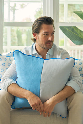 A man with light brown hair and a beard sits on a sofa holding a Spring Blue Blocks Oversized Pillow, adding a modern, calming vibe. He wears a light blue shirt and beige pants; patterned pillows and a green plant are in the background.