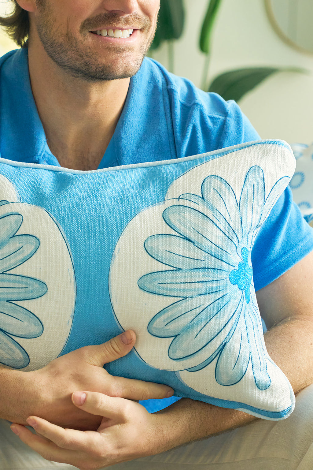 A man in a blue shirt hugs the Calico Scallop Lumbar Pillow, featuring white and blue floral designs. Only his smiling mouth and hands are visible, evoking a sophisticated, coastal summer vibe.