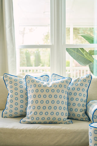 A beige sofa with three white pillows—including a Calico Mini Floral Oversized Pillow and two with bright blue piping—rests before a sunlit window and green plant in this cozy, inviting room.