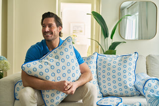 A man sits on a white couch, smiling and hugging a Calico Mini Floral Oversized Pillow with bright blue piping, surrounded by matching cushions in a bright, modern living room.