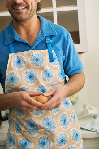 A person in a blue shirt smiles while holding an egg in their kitchen. They wear the Calico Scallops on Yellow Apron, reminiscent of Sewing Down South style, with large blue flowers and an adjustable neck strap. White cabinets provide a crisp backdrop.
