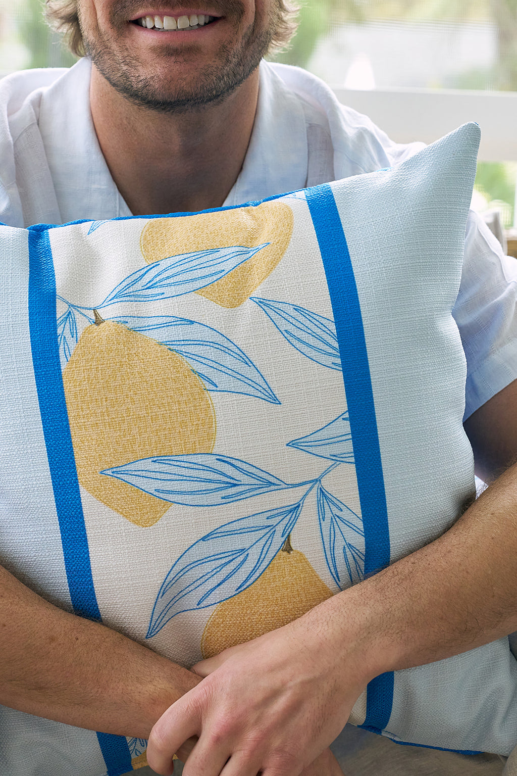 Two Lemons in a Line Pillows with a yellow lemon and blue leaf design sit on a beige wicker outdoor sofa. A green plant and white railing complete this cozy, sunlit patio, ideal for citrus-themed decor.