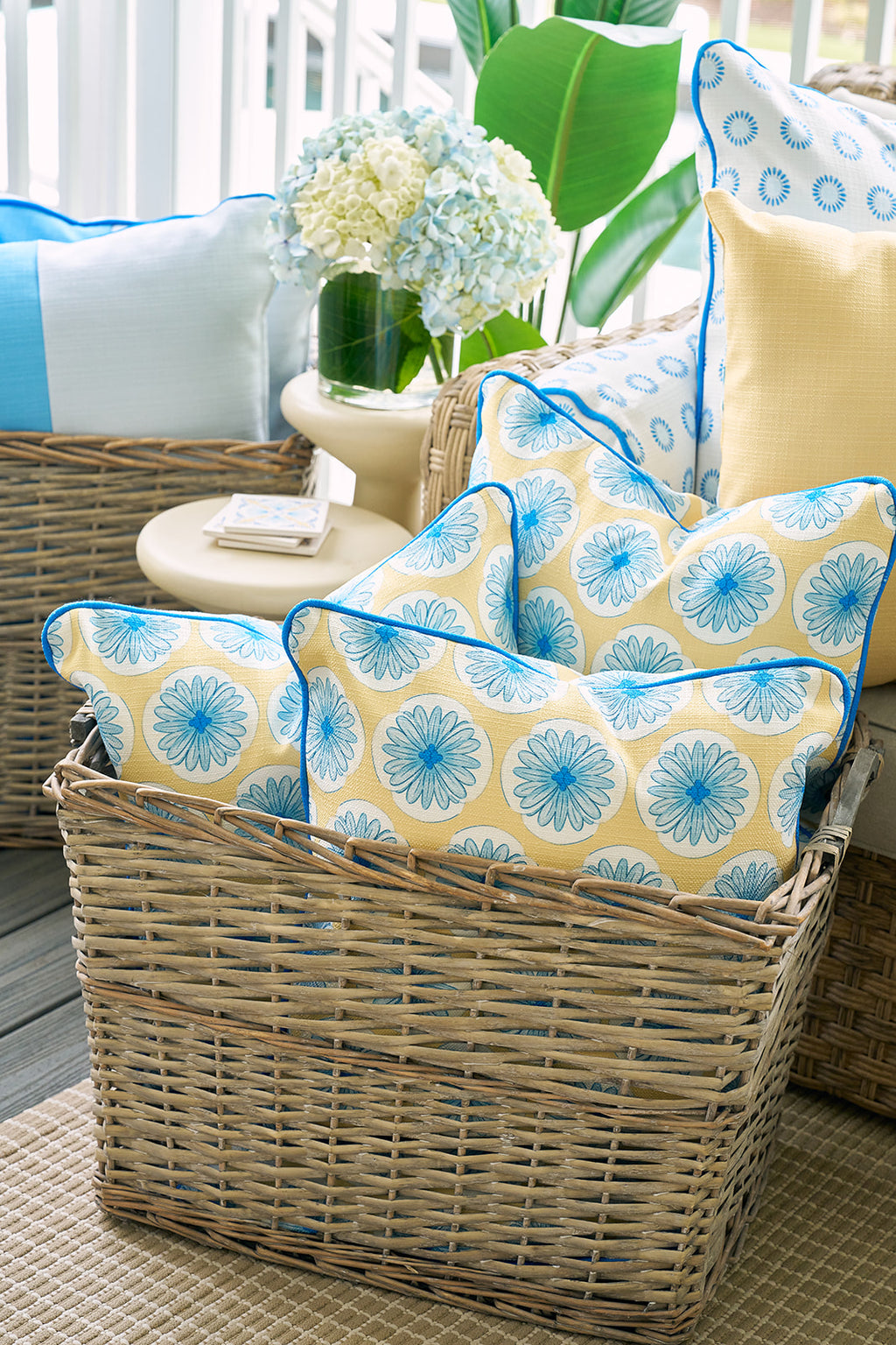 The Yellow Calico Scallops Pillow, with blue floral patterns and piping, creates a sophisticated accent on a beige sofa. In the blurred background, a person in a blue shirt stands holding a cup.
