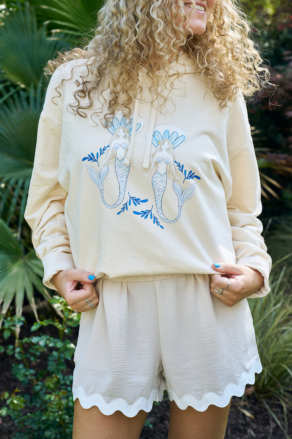 Two women relax outdoors on a lounge chair, smiling and wearing pastel SDS Sirens Hoodies from The Siren Collection. A beige tote with a blue mermaid print rests nearby amid lush greenery and coastal vibes.