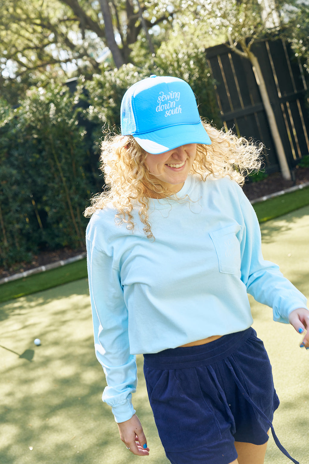 A man stands poolside, smiling and holding the brim of a Sewing Down South Trucker from The Siren Collection in coastal blue. He wears a light t-shirt and beige pants, surrounded by greenery and trees.