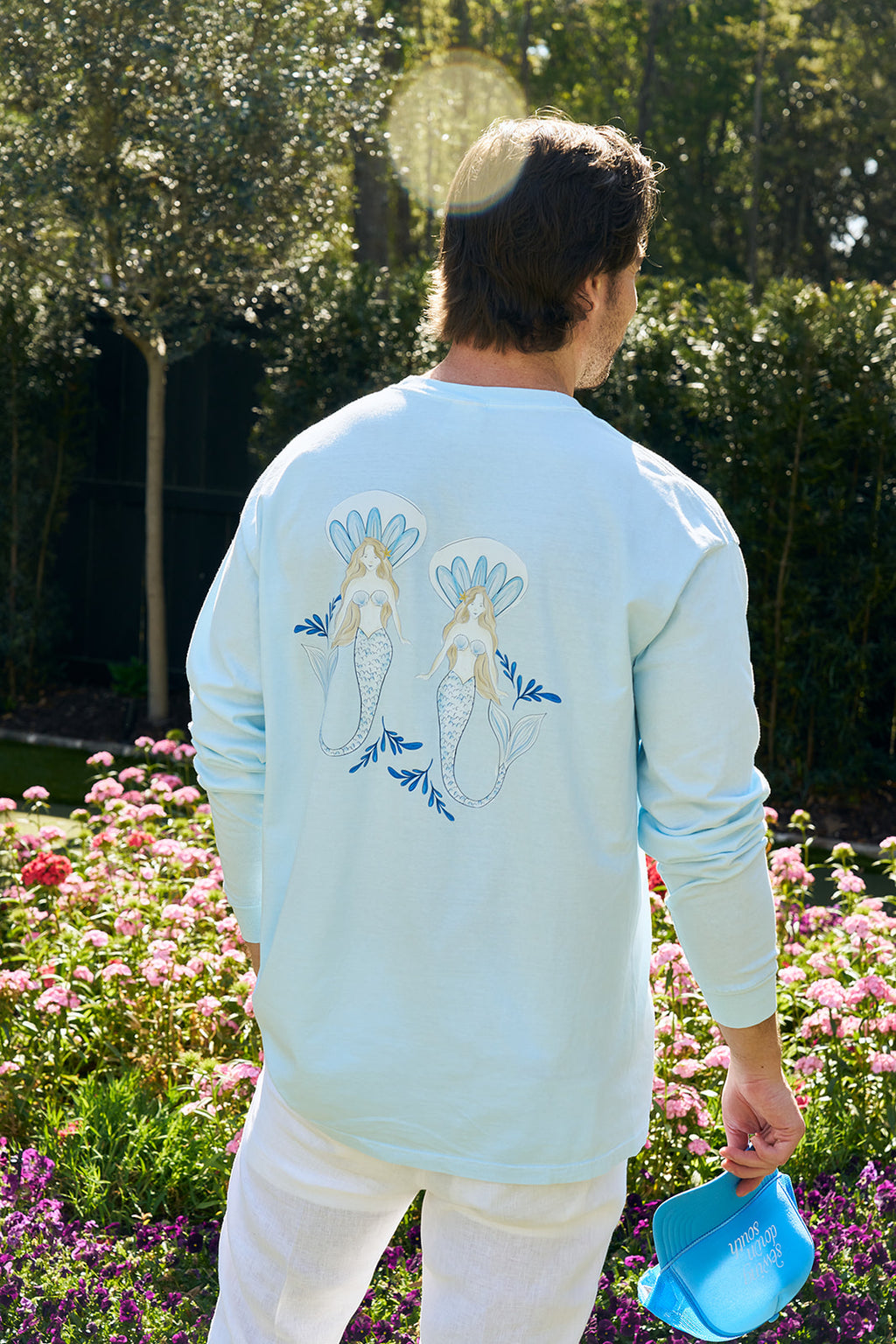 Dressed in The Siren Collections heavyweight, unisex relaxed fit long sleeve tee and white pants, a person smiles amidst a garden filled with pink flowers. Sunlight filters through the trees in the background, crafting a serene atmosphere.