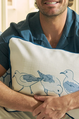 A person wearing a blue shirt holds a Sandpiper Trio with Blue Lattice Lumbar Pillow. Only the lower half of their face and upper torso are visible, with a softly lit, out-of-focus background.