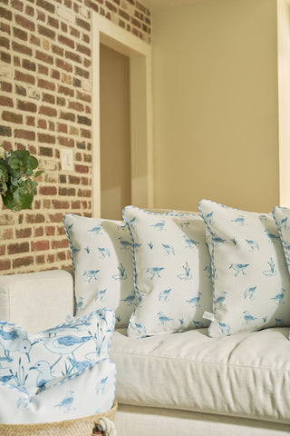 A white couch with four light cushions, including The Sandpiper Pillow featuring blue bird and plant designs, sits against a brick wall in a cozy living room. A basket with a matching cushion rests on the armrest.