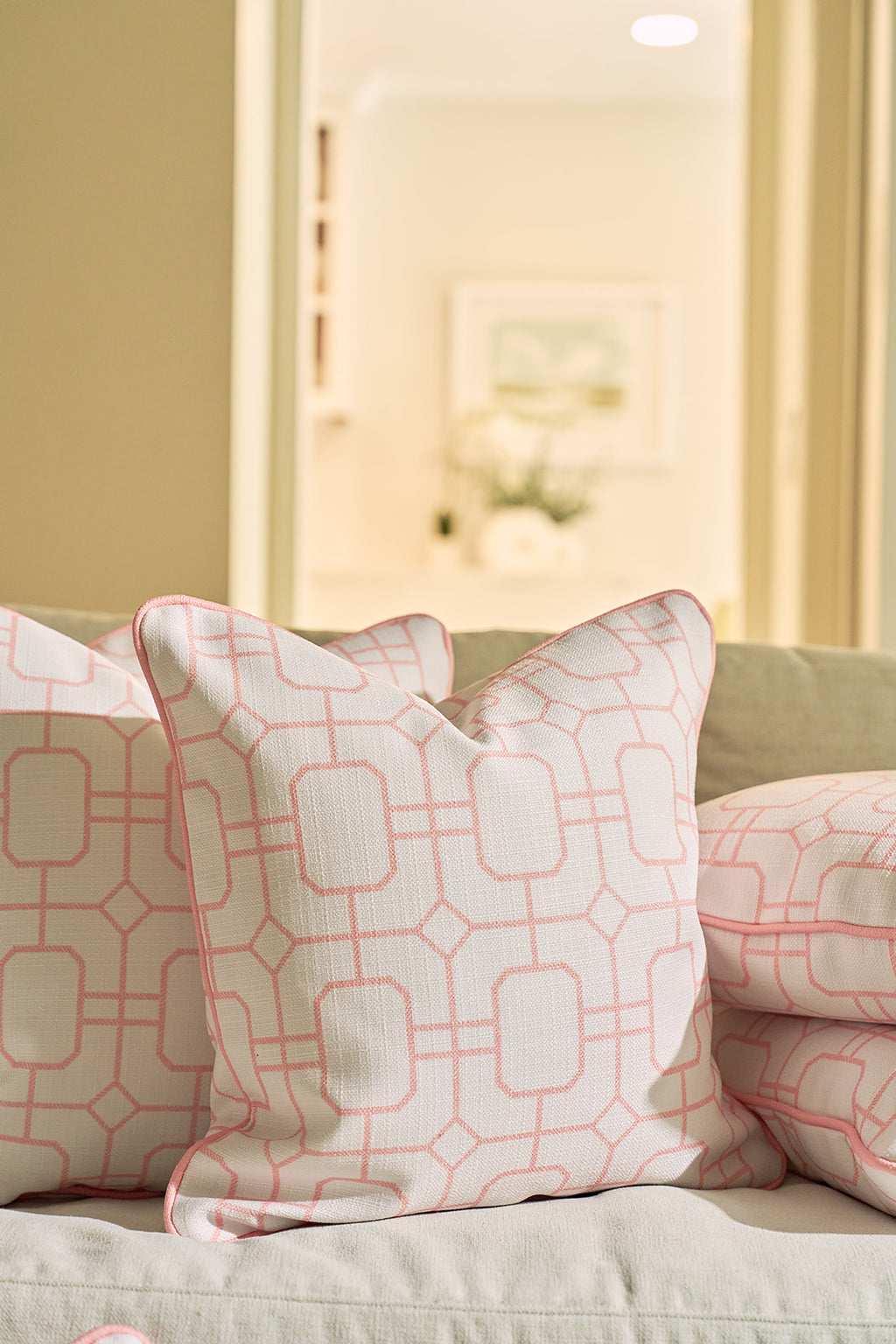 A smiling man with light brown hair and a beard, in a pale pink shirt and white pants, sits on a light sofa holding the Pink Lattice Pillow, which features timeless charm and a classic geometric lattice design.