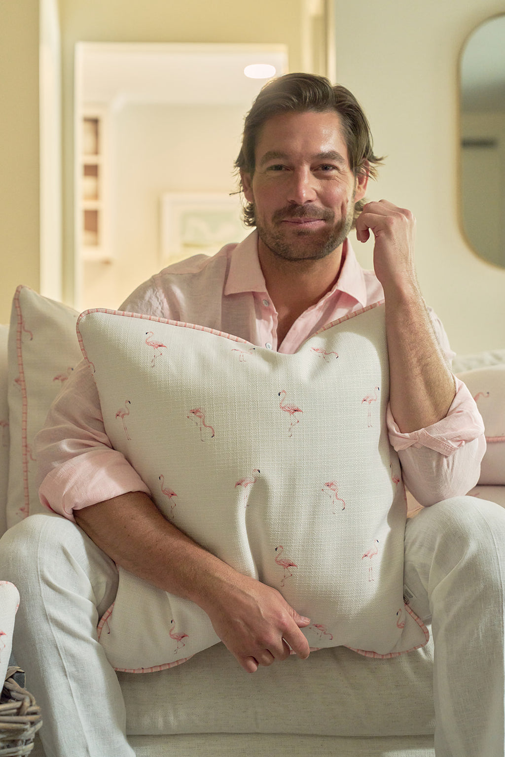 A person in a light pink shirt smiles while holding the Flamingo Scatter Oversized Pillow, featuring small pink flamingos on durable fabric. Another pillow with an elevated twisted welt is seen in the background.