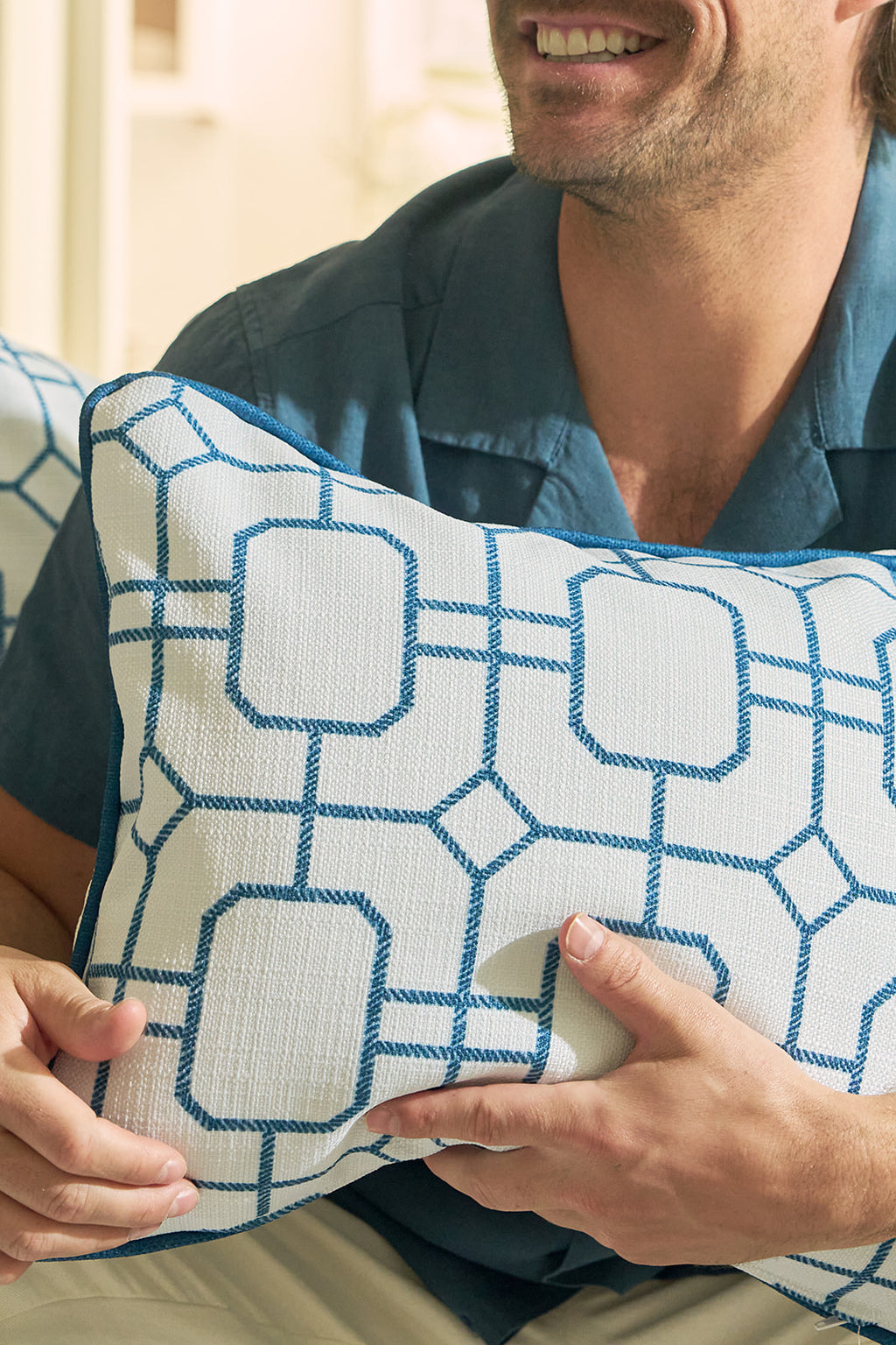 A man sits on a cream sofa, smiling and giving a thumbs up while holding a Sandpiper Trio with Blue Lattice Lumbar Pillow. The sofas blue and white patterned pillows enhance the cozy, beachy vibe of the neutral room.