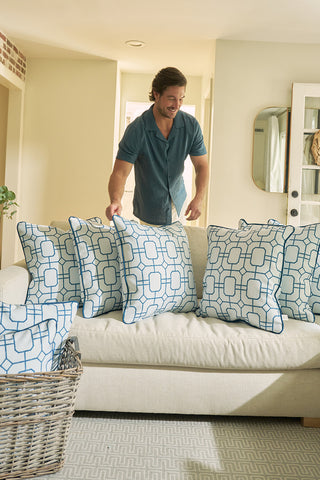A man in a blue shirt arranges a Blue Lattice Oversized Pillow and other patterned pillows on a cream sofa in a bright, cozy living room with light walls and a pillow-filled basket, creating fresh coastal décor.