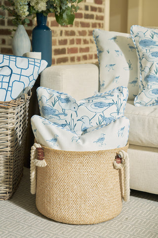 A woven basket with rope handles holds two Sandpiper Pillows in blue and white coastal motifs beside a light sofa with matching pillows. Nearby, a wicker basket and blue vase rest against a brick wall.